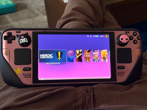 Pink steamdeck with a black protective case around it. There are three stickers on it. A flower, a star, and a heart. There are two silicon thumbstick covers. One is kuromi from hello kitty and the other is a cute pink skull. It is the image of peak gaming. 