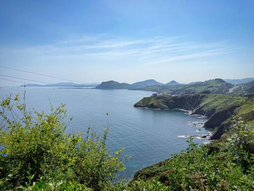 Paesaggio costiero con mare, scogliere verdi e colline in lontananza sotto un cielo azzurro con alcune nuvole.